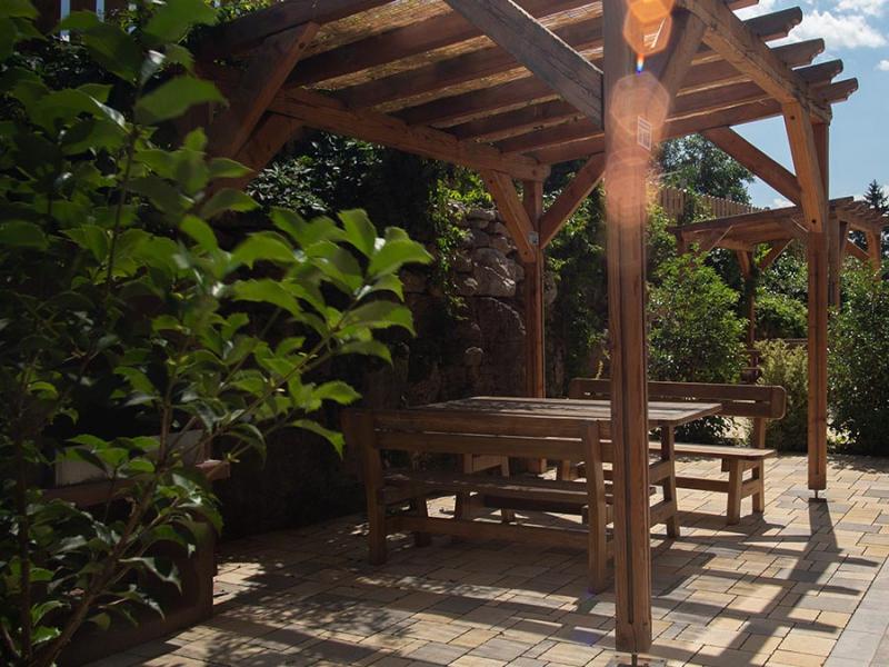 Pergolato in legno con tavolo e panchine, immerso in un giardino soleggiato.